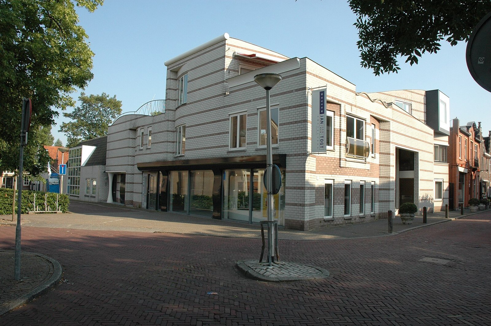 A light-striped brick museum building on a cobbled street corner, with large shop windows on the ground floor and a vertical name sign on the façade.
