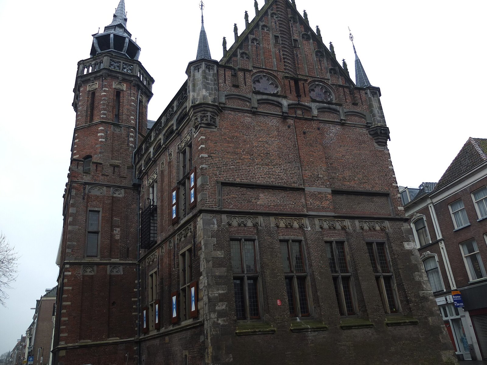 Late Gothic brick town hall with a tall tower with an open lantern on the left, a pointed gable with pinnacles and turrets, and below, large leaded-glass windows with red, white and blue shutters, seen from the street.