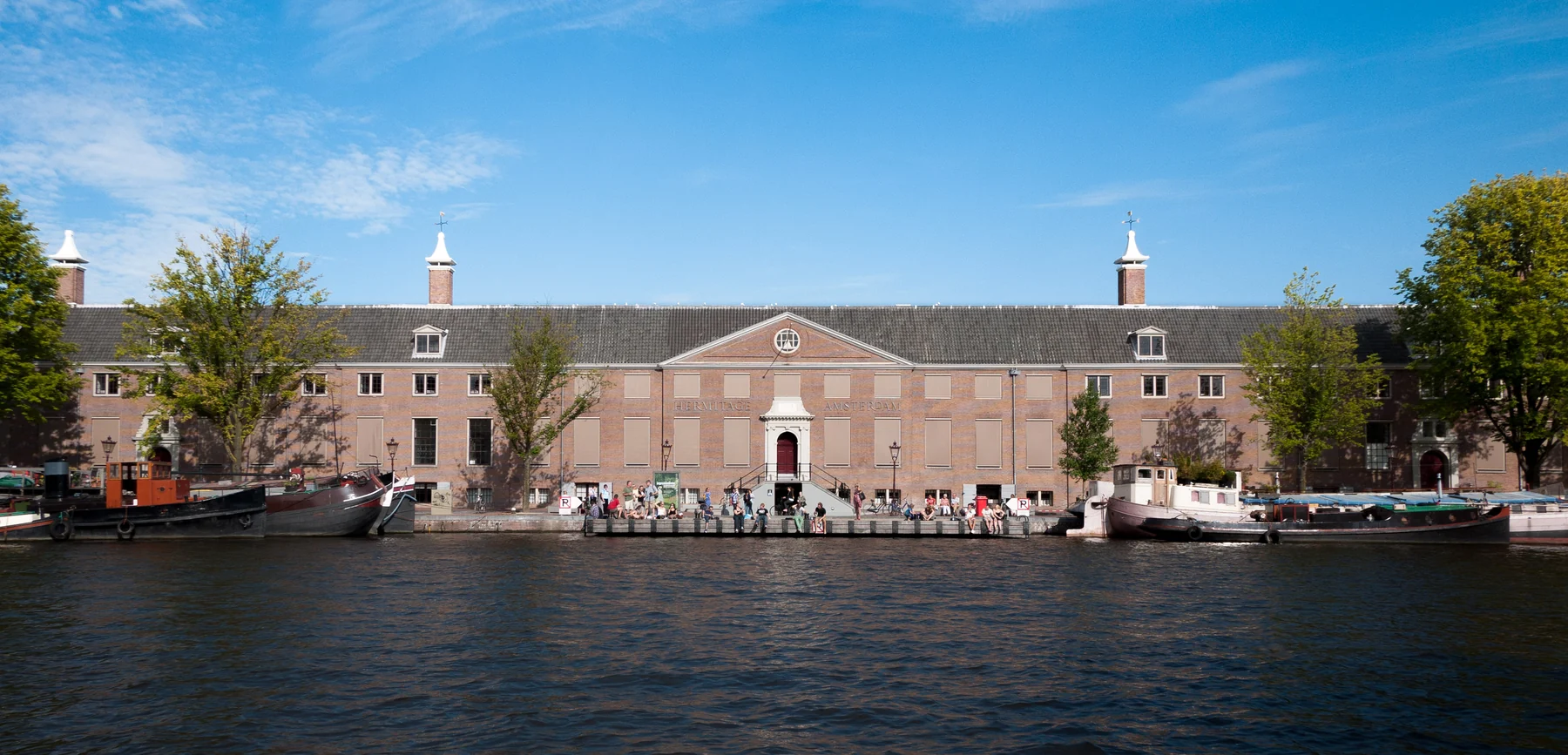 Lange Backsteinfassade des Museums an der Amstel, mit Booten und Besuchern am Kai.