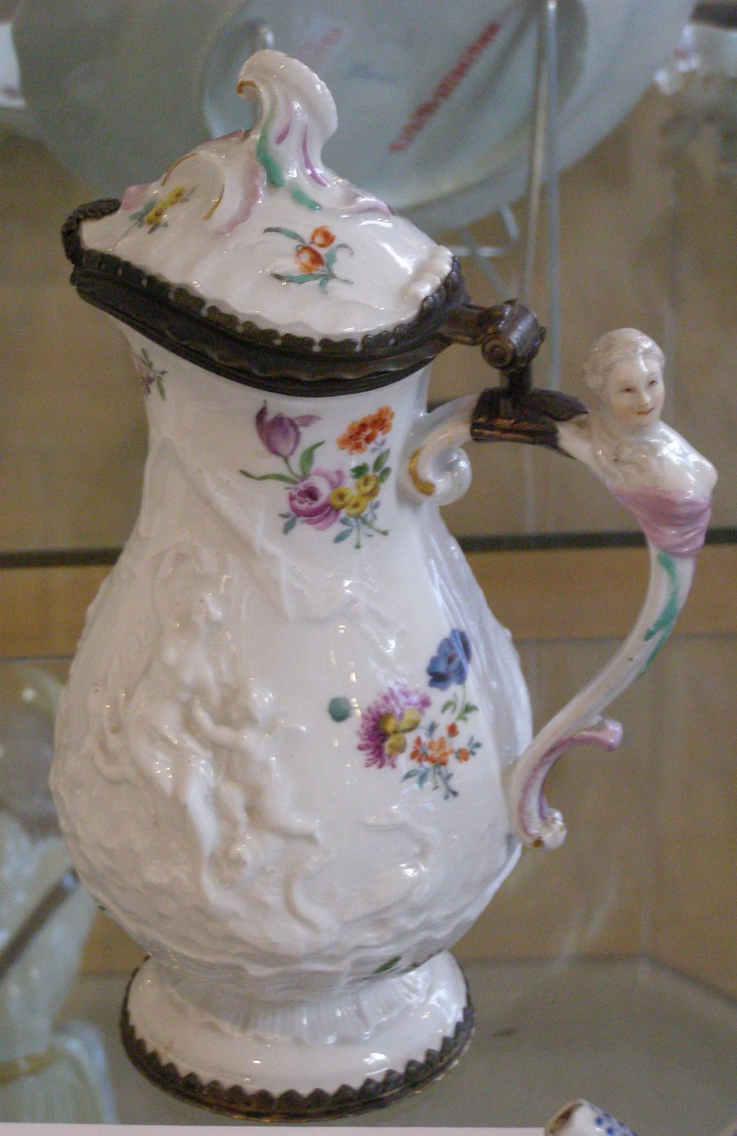 Witte koffiekan met bloemen, reliëfdecoratie en een mensfiguur op het handvat.