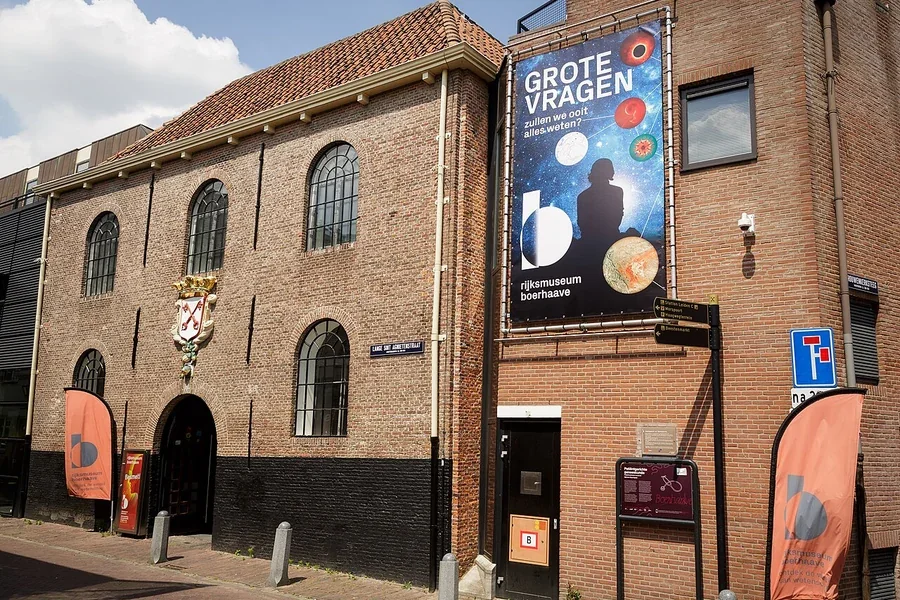 Bakstenen gevel en boogvormige ingang van Rijksmuseum Boerhaave in Leiden, met een grote blauwe poster aan de muur.