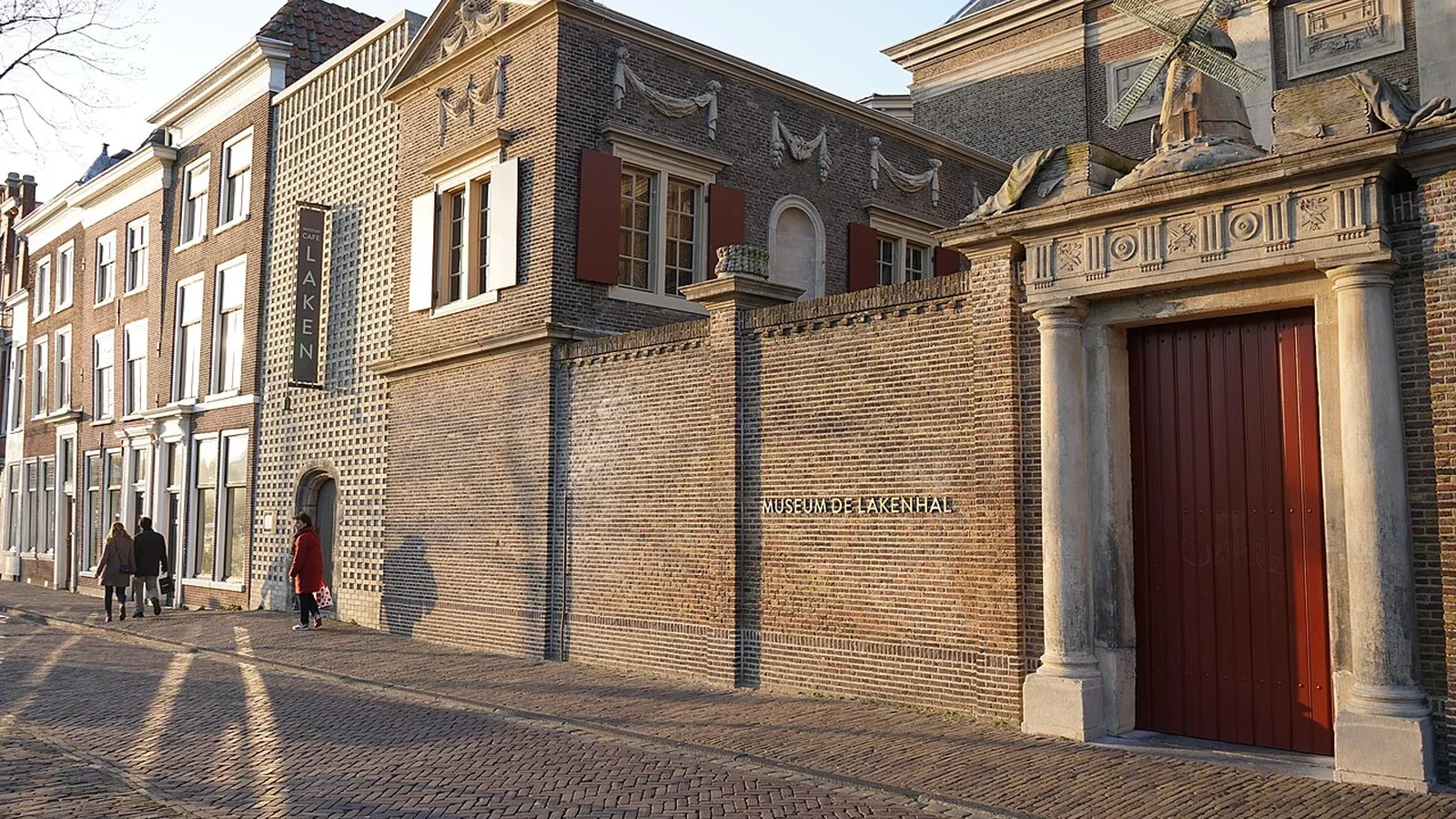 Bakstenen gevel van Museum De Lakenhal aan een straat in Leiden, met een grote rode deur en enkele voorbijgangers.