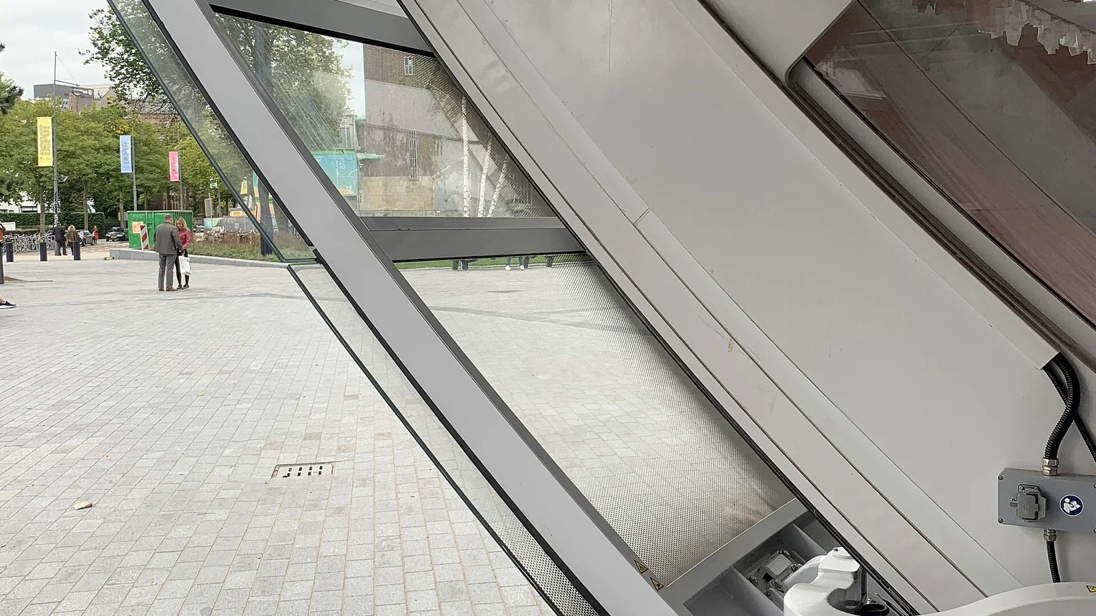Schräge Konstruktion aus Glas und Metall am Depoteingang mit Blick auf den Museumpark und Besucher draußen.