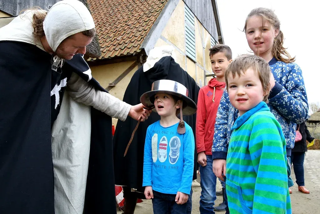 Historische bewoner laat kinderen een metalen helm passen bij een gebouw in het voormalige preHistorisch Dorp.