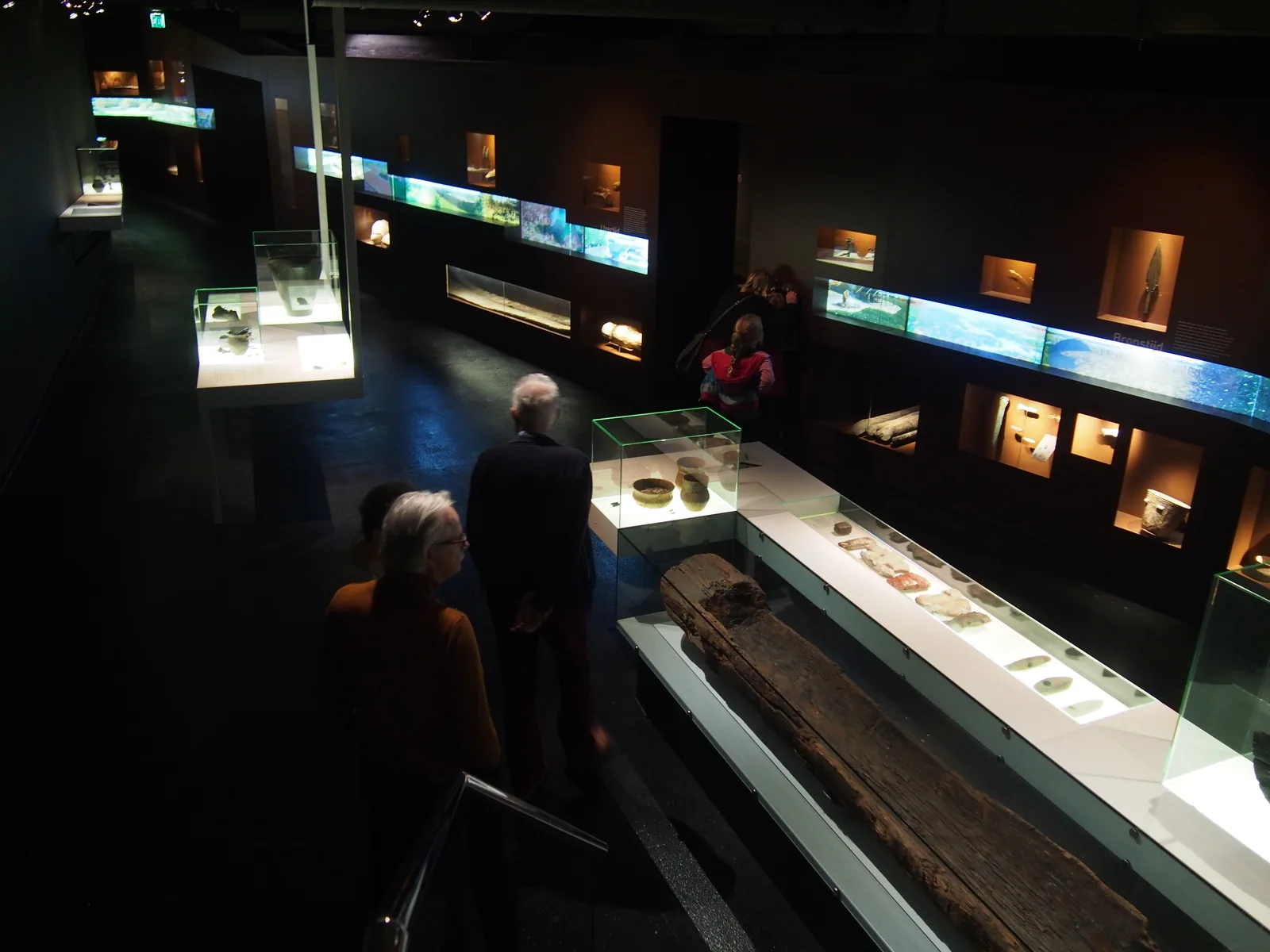 Donkere museumzaal met bezoekers en de lange houten kano van Pesse in een verlichte vitrine.