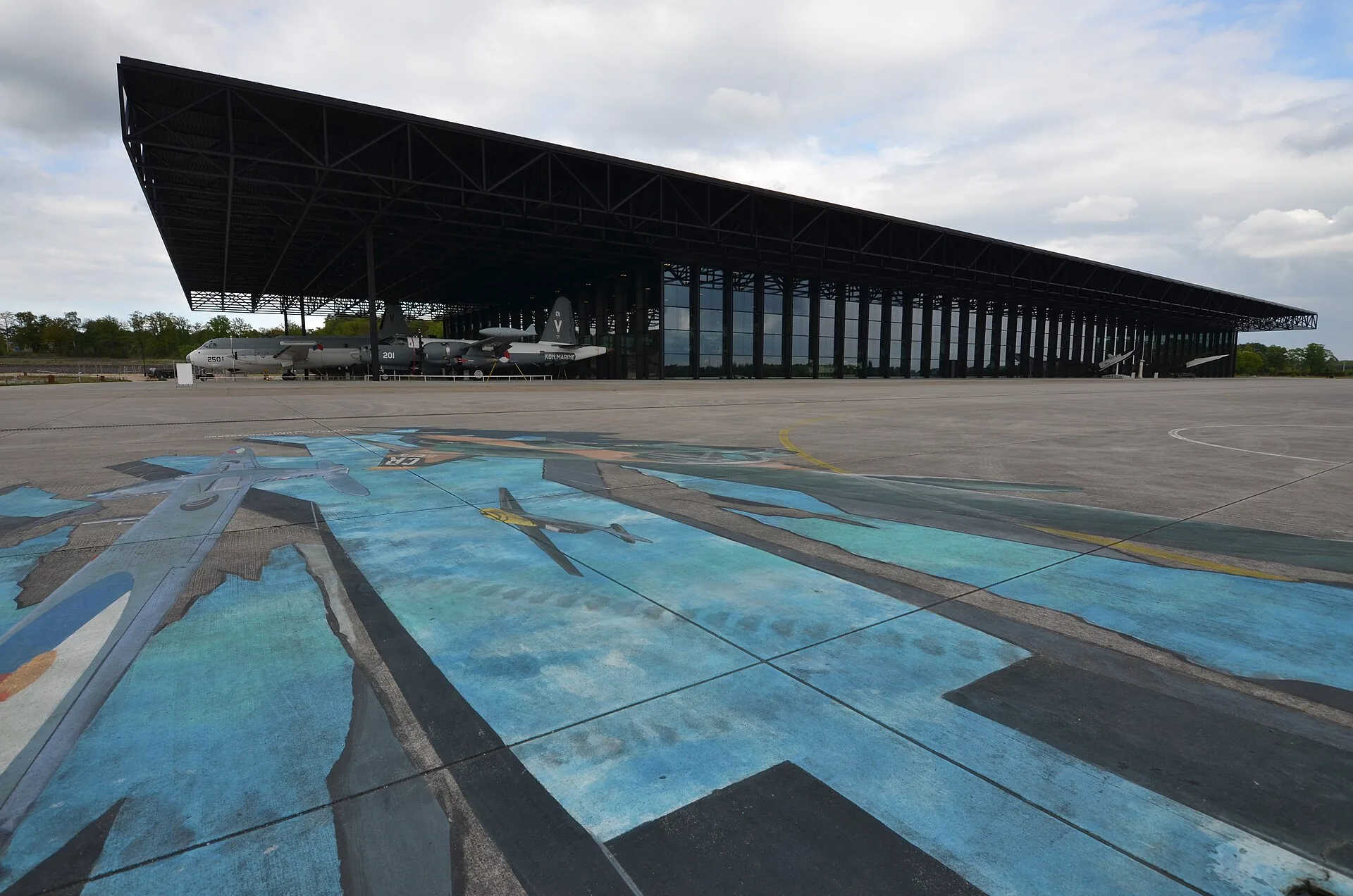 Het brede museumgebouw met vliegtuigen onder de luifel en een geschilderd platform op de voorgrond.
