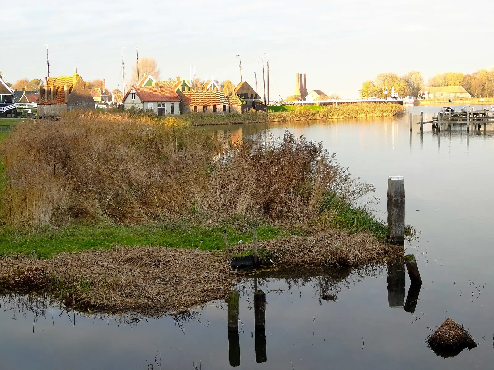 Huizen met pannendaken aan een brede waterpartij, met riet en scheepsmasten op de achtergrond.