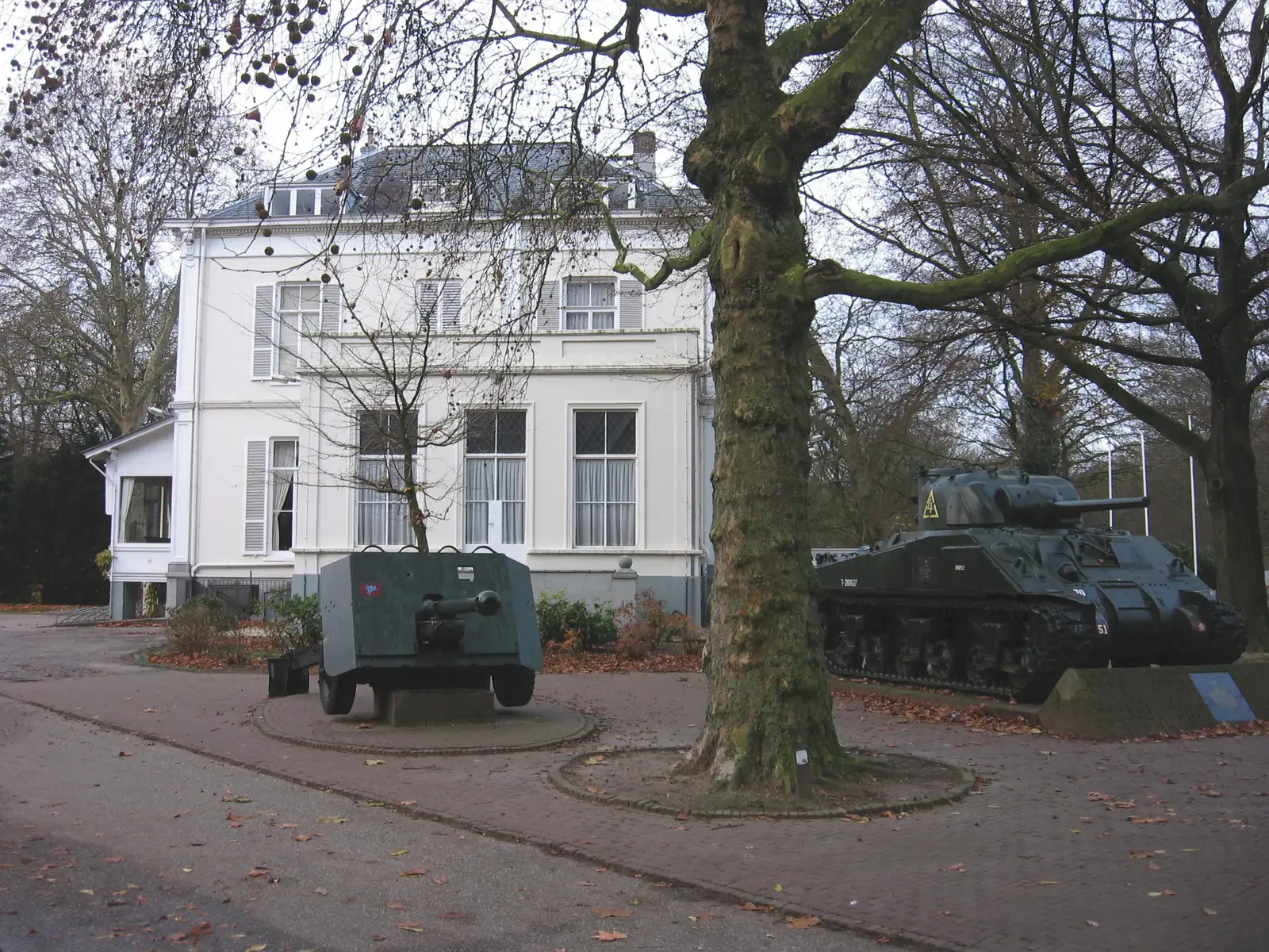 Vue ancienne de la villa Hartenstein avec des objets militaires dehors et un arbre au premier plan.